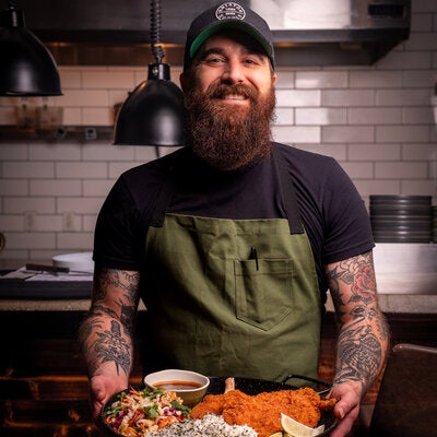 Smiling bearded man in a green apron holds a platter of breaded cutlets, rice, slaw, lime wedges, and sauce in a tiled kitchen.